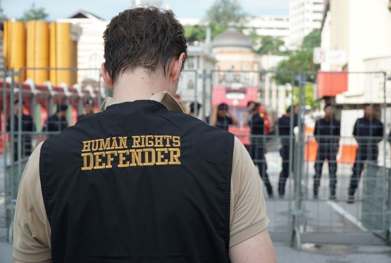 A man with a gilet from the back, with Human Rights Defender written on it